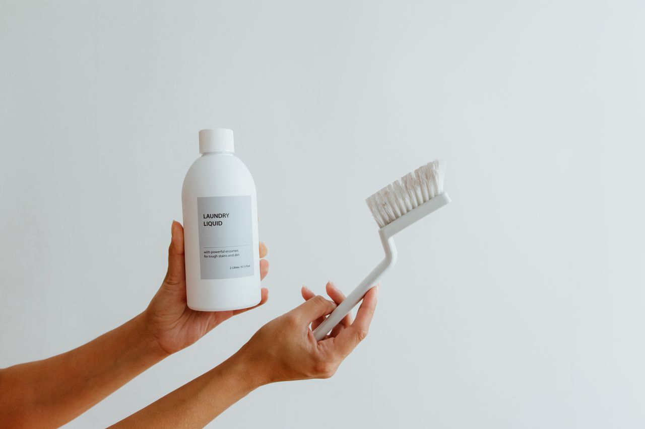 creative Close-up of hands holding a laundry liquid bottle and cleaning brush against a white background.
