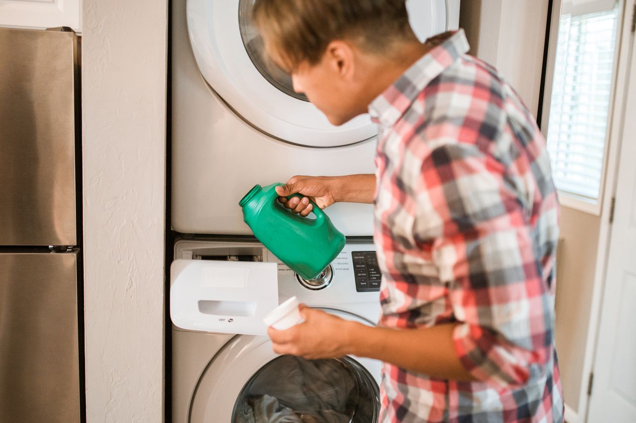 Services-01 A man in a plaid shirt pours detergent into a front-loading washing machine, engaging in a household chore.